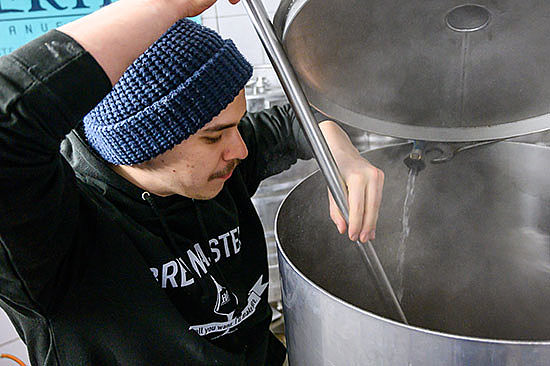 Ein junger Mann mit Mütze rührt mit einem langen Stab in einem großen Tank, aus dem es dampft.  