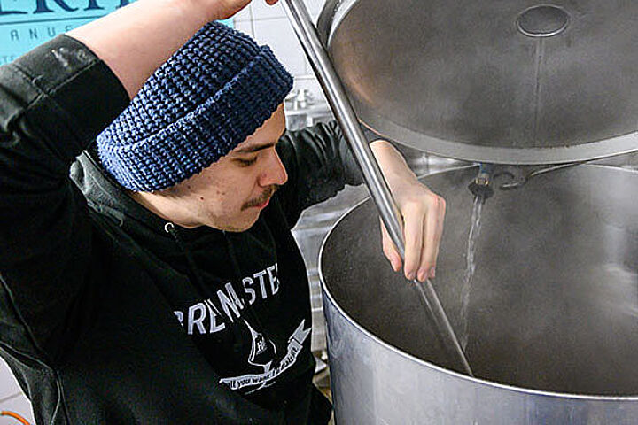 Ein junger Mann mit Mütze rührt mit einem langen Stab in einem großen Tank, aus dem es dampft.  