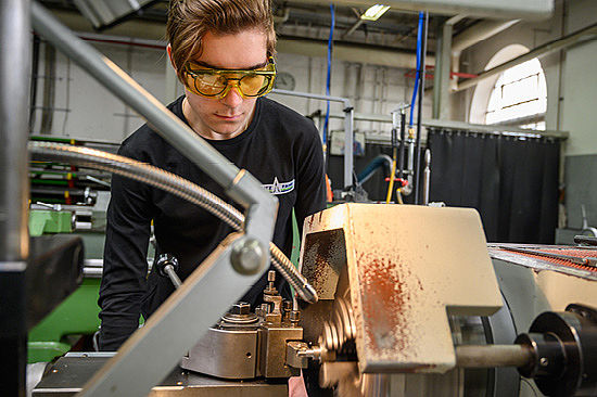 Ein junger Mann mit Schutzbrille steht vor einer Drehmaschine. 
