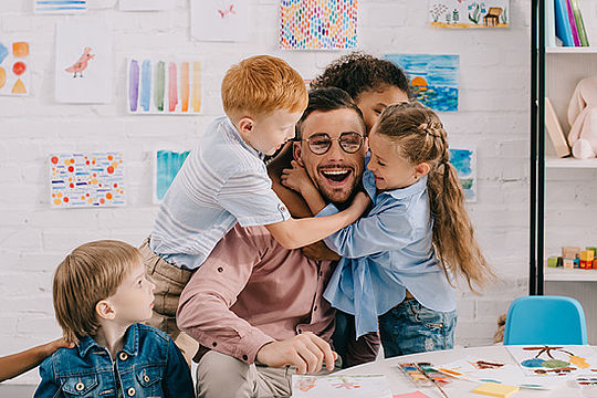 Ein lachender Mann sitzt an einem Tisch mit Malsachen. Lachende Kinder fallen ihm um den Hals.