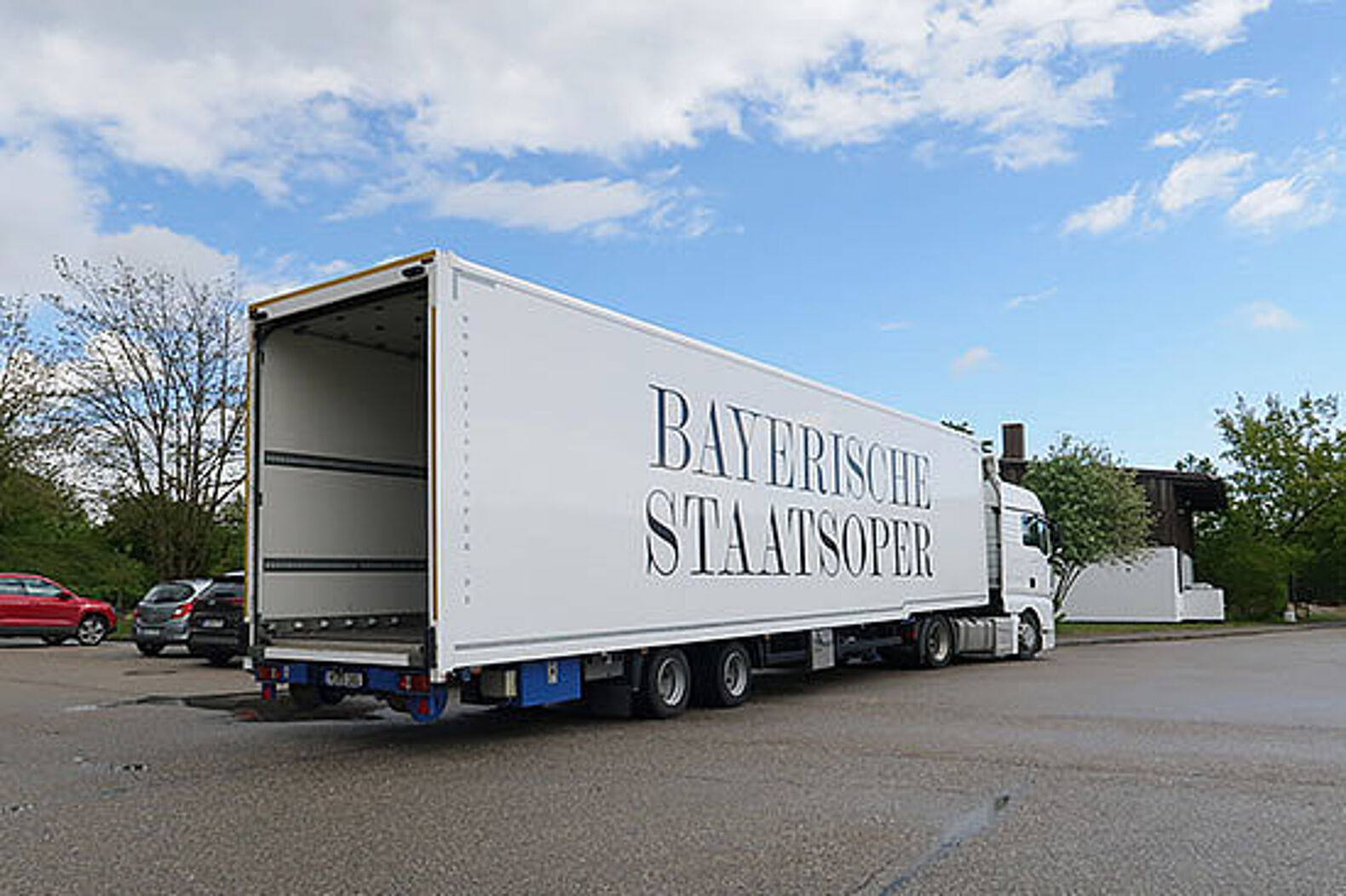 Auf einem Parkplatz: Ein großer LKW mit der Aufschrift „Bayerische Staatsoper“ wartet mit geöffneter Heckklappe aufs Beladen.