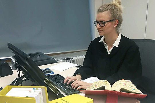 Justizfachwirtin Julia bei der Arbeit am Computer. Auf ihrem Schreibtisch liegen mehrere Gesetzesbücher.