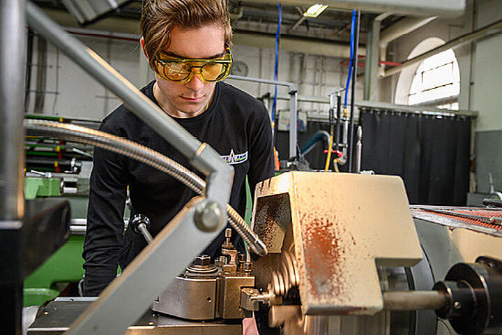 Ein junger Mann mit Schutzbrille steht vor einer Drehmaschine. 