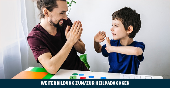 Ein Mann mit Bart und zusammengebundenen Haaren faltet seine Hände und schaut einen Jungen an. Der Junge ahmt ihn nach und lächelt dabei.