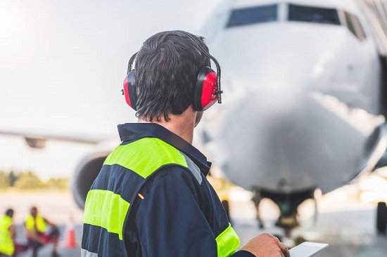 Ein Mann mit roten Ohrenschützern, gelber Weste und einem Tablet steht vor einem weißen Flugzeug, zwei Männer mit gelben Westen Knien links neben dem Flugzeug.