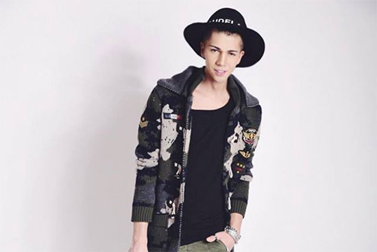 Designer Maximilian Seitz trägt einen schwarzen Hut, eine offene dunkelgraue Jacke mit hellen Prints, ein schwarzes T-Shirt und eine grüne Hose, die rechte Hand hängt am Körper und die linke Hand ist in der Hosentasche.