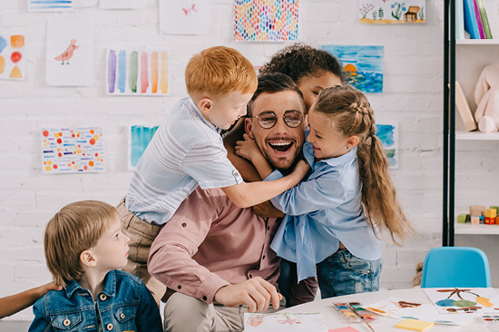 Ein lachender Mann sitzt an einem Tisch mit Malsachen. Lachende Kinder fallen ihm um den Hals.  