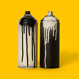 Vor gelbem Hintergrund stehen zwei Sprühflaschen: eine graue mit heruntergelaufener schwarzer Farbe und eine schwarze mit heruntergelaufener grauer Farbe.