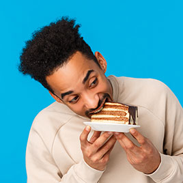 Ein Mann steht vor blauem Hintergrund und beißt in ein großes Stück Torte.